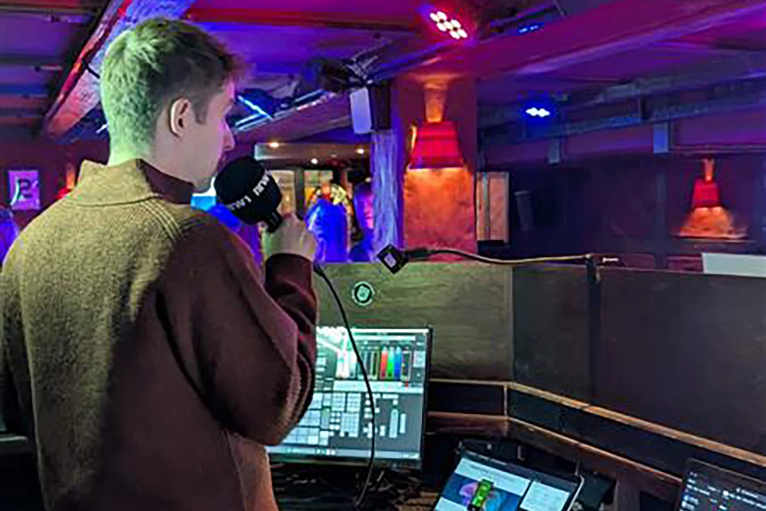 Person mit Mikrofon stehend an einem Technikpult hält Vortrag.