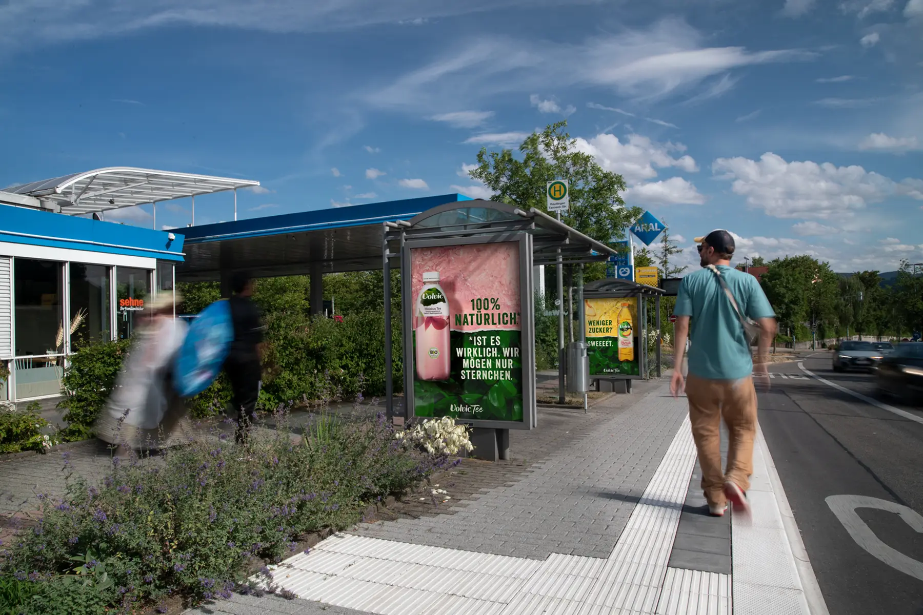 Man walks on sidewalk past bus stops with drink ads under a sunny sky.
