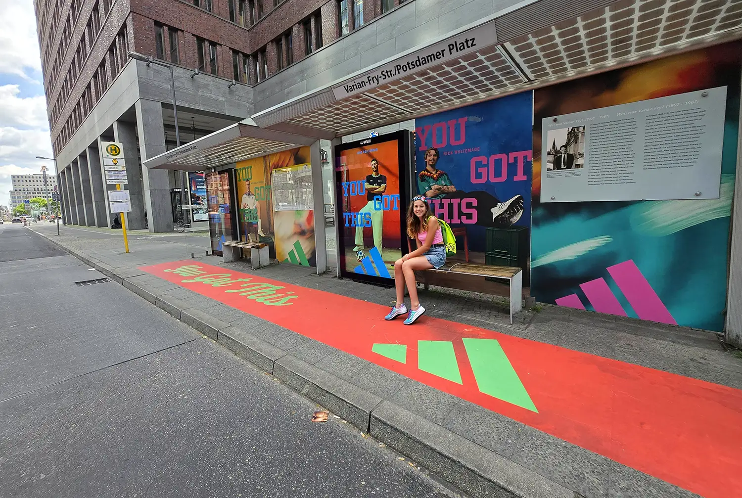 A bus stop with Adidas "YOU GOT THIS" ads, a woman sitting on a bench, and red pavement.
