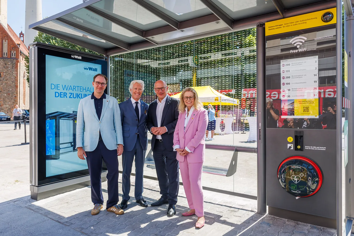 Vier Personen stehen vor einer modernen Wartehalle mit Digital City Light Board und Defibrilator.