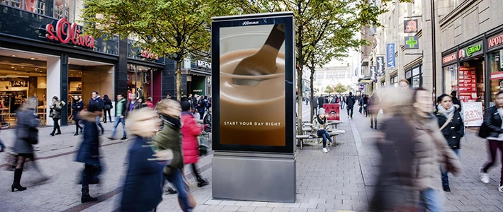 Digital City Light Poster mit Kaffee-Werbung in einer belebten Einkaufsstraße in Hamburg, umgeben von Geschäften und vorbeigehenden Menschen.