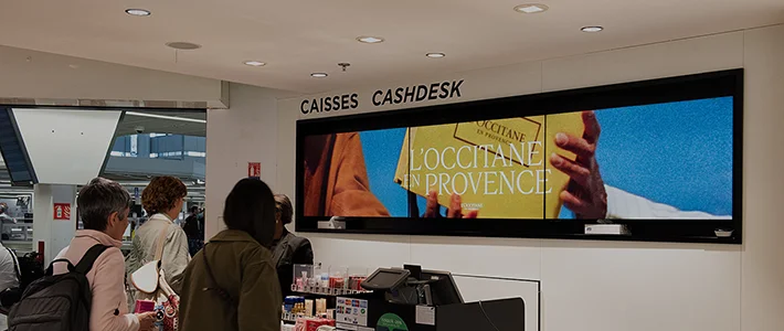 Digitaler Werbeträger hinter einem Counter am Flughafen in Paris zeigt Kosmetikwerbung.